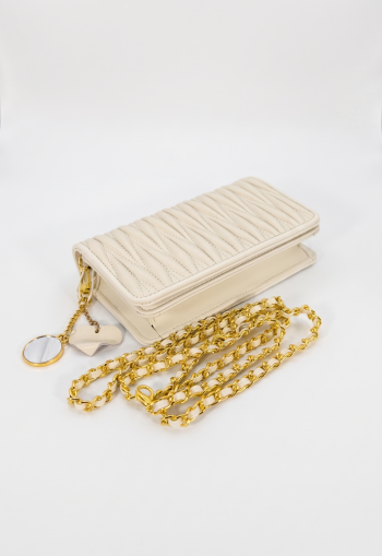 Off-white matelassé textured leather crossbody bag by SHUCLUB, featuring a distinctive vertical quilted pattern, polished gold-tone hardware, a woven leather chain strap, and a decorative heart and mirror charm, shot on a clean white background.