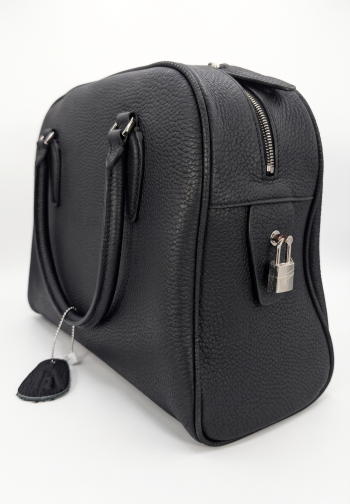 Side Profile View: Onyx black pebbled leather doctor-style satchel bag by SHUCLUB, showcasing a structured dome silhouette, rolled leather top handles, a silver-tone zipper, and a decorative silver padlock attached to a side leather tab, shot on a white background. Front View: Full front studio view of the black pebbled leather SHUCLUB handbag highlighting its rich grain texture, clean stitched seams, arched top handles with silver metal connectors, and minimalist profile.