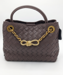 Front View: Chocolate brown woven leather tote bag by SHUCLUB, featuring a classic basketweave intrecciato pattern, a structured top rolled handle, large gold-tone grommets, and an ornamental chunky gold chain with a central infinity knot detail, shot on a solid white background. Top Angle View: Top-down studio shot of the brown woven SHUCLUB tote bag showcasing the dual open compartments, internal magnetic tab closure, and tucked-in leather top handle with draped gold hardware accents.